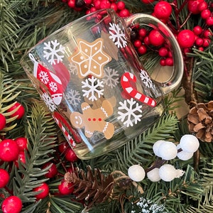 May include: A clear glass mug with a white handle decorated with festive Christmas designs. The mug features snowflakes, candy canes, gingerbread men, and a gingerbread star. The mug is surrounded by a green Christmas wreath with red berries.