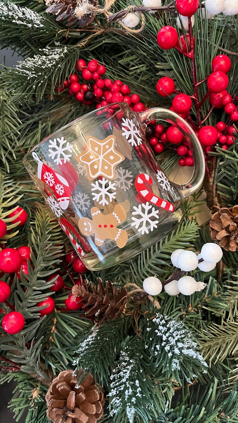 May include: A clear glass mug with a white handle decorated with festive Christmas designs. The mug features snowflakes, candy canes, gingerbread men, and a gingerbread star. The mug is surrounded by a green Christmas wreath with red berries.