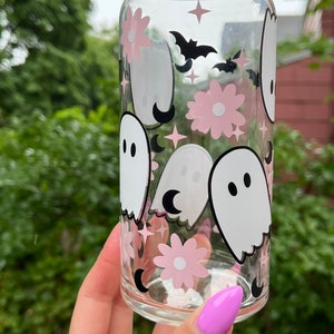 May include: A clear glass can with a pink and white ghost and flower pattern. The can has a black bat and crescent moon design.