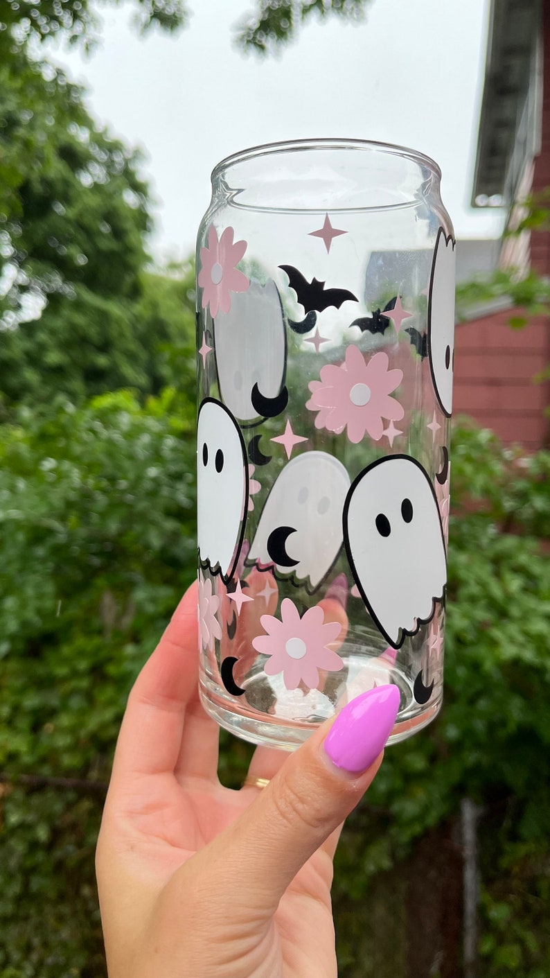 May include: A clear glass can with a pink and white ghost and flower pattern. The can has a black bat and crescent moon design.