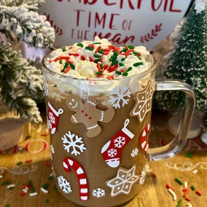 May include: A clear glass mug with a brown handle, decorated with Christmas-themed stickers, filled with a brown beverage topped with whipped cream and red and green sprinkles. The mug is sitting on a wooden surface with more sprinkles scattered around it. The background features a white sign with the text "Wonderful Time of the Year" in red.