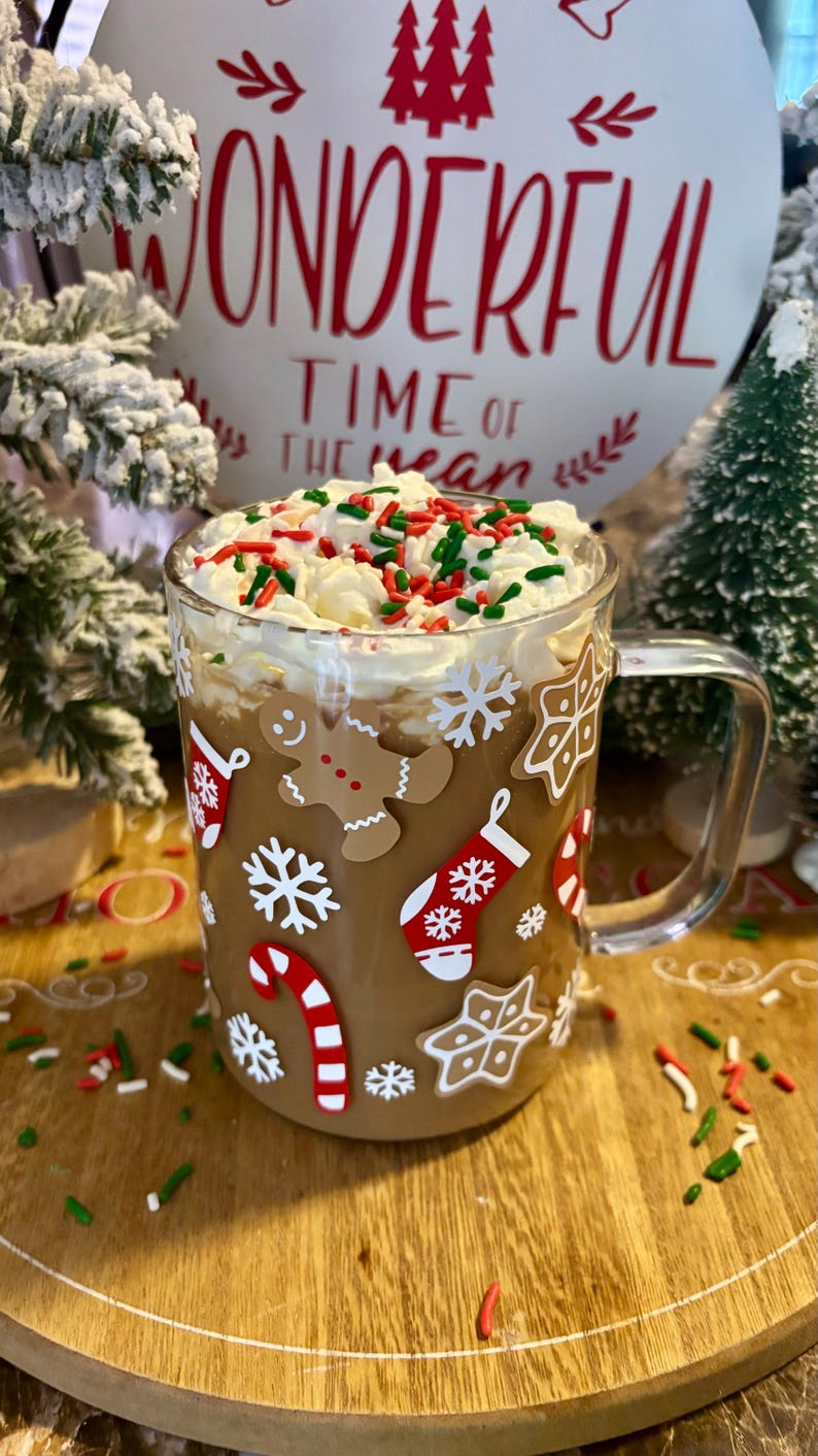 May include: A clear glass mug with a brown handle, decorated with Christmas-themed stickers, filled with a brown beverage topped with whipped cream and red and green sprinkles. The mug is sitting on a wooden surface with more sprinkles scattered around it. The background features a white sign with the text "Wonderful Time of the Year" in red.