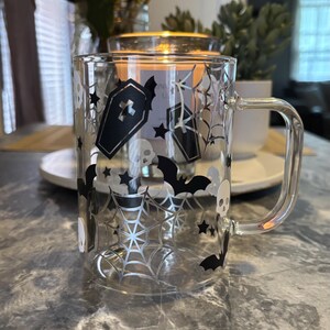 Pode incluir: Uma caneca de vidro transparente com um design de Halloween em preto e branco. O design apresenta morcegos, teias de aranha, caix&otilde;es e estrelas. A caneca tem uma al&ccedil;a.