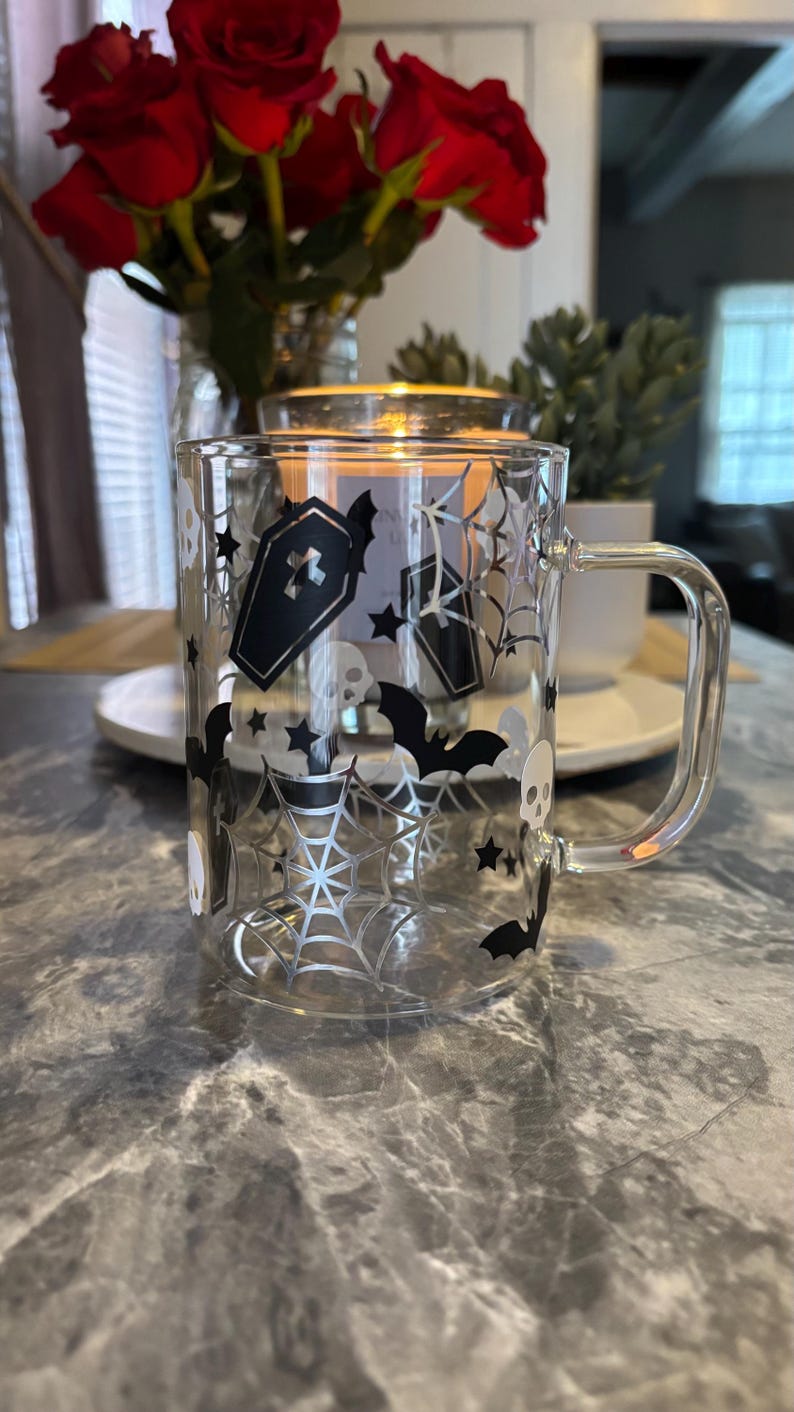 Pode incluir: Uma caneca de vidro transparente com um design de Halloween em preto e branco. O design apresenta morcegos, teias de aranha, caix&otilde;es e estrelas. A caneca tem uma al&ccedil;a.
