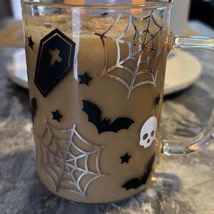 Pode incluir: Uma caneca de vidro transparente com um design de Halloween preto e prata. A caneca est&aacute; cheia de um l&iacute;quido castanho e tem uma pega. O design inclui um caix&atilde;o, morcegos, estrelas e teias de aranha.