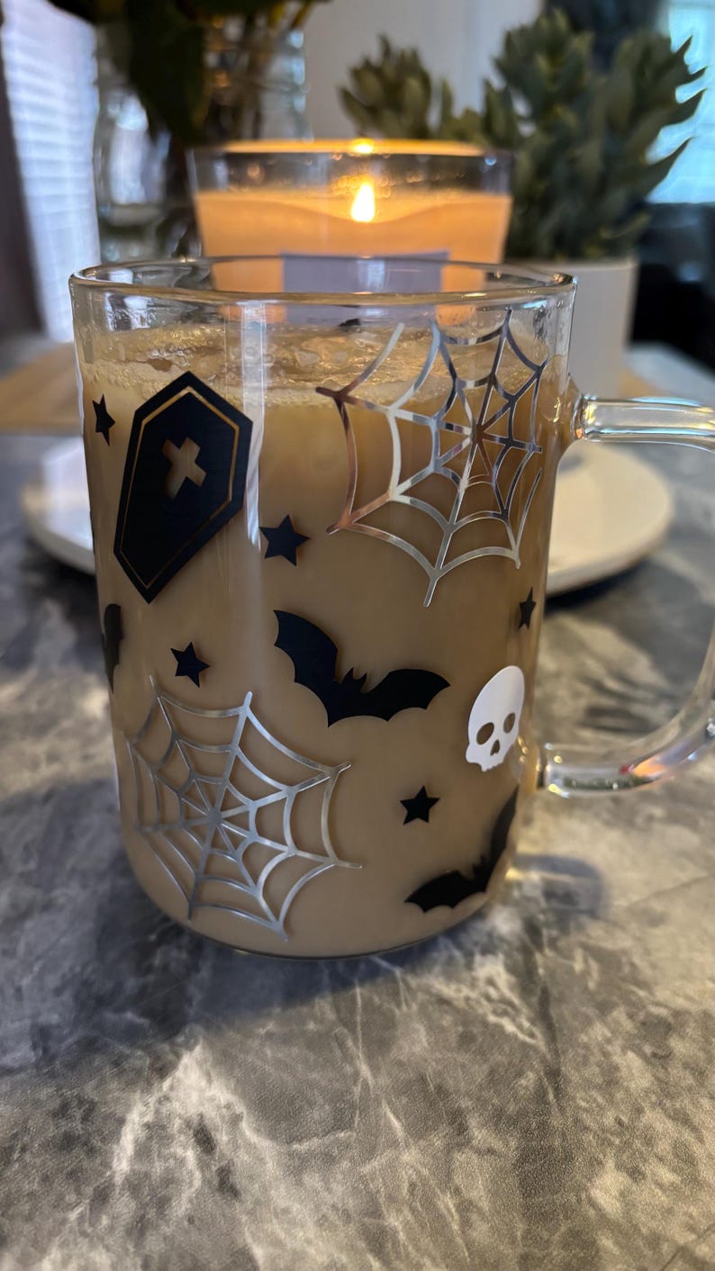 Pode incluir: Uma caneca de vidro transparente com um design de Halloween preto e prata. A caneca est&aacute; cheia de um l&iacute;quido castanho e tem uma pega. O design inclui um caix&atilde;o, morcegos, estrelas e teias de aranha.