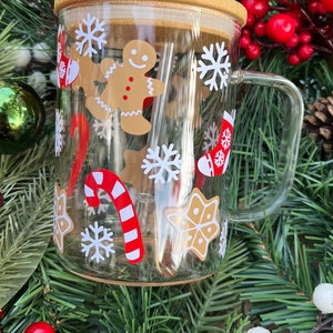 May include: A clear glass mug with a brown lid and a red and white striped straw. The mug has a Christmas theme with gingerbread men, candy canes, snowflakes, and stockings.