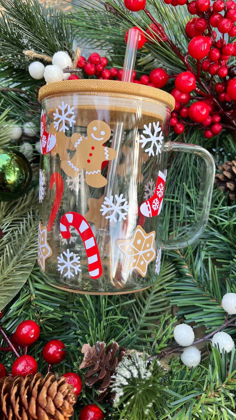 May include: A clear glass mug with a brown lid and a red and white striped straw. The mug has a Christmas theme with gingerbread men, candy canes, snowflakes, and stockings.