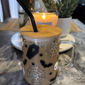 Pode incluir: Uma caneca de vidro com um design de Halloween marrom e preto. A caneca tem uma tampa de bambu e um canudo preto. O design inclui morcegos, teias de aranha, estrelas e caveiras.