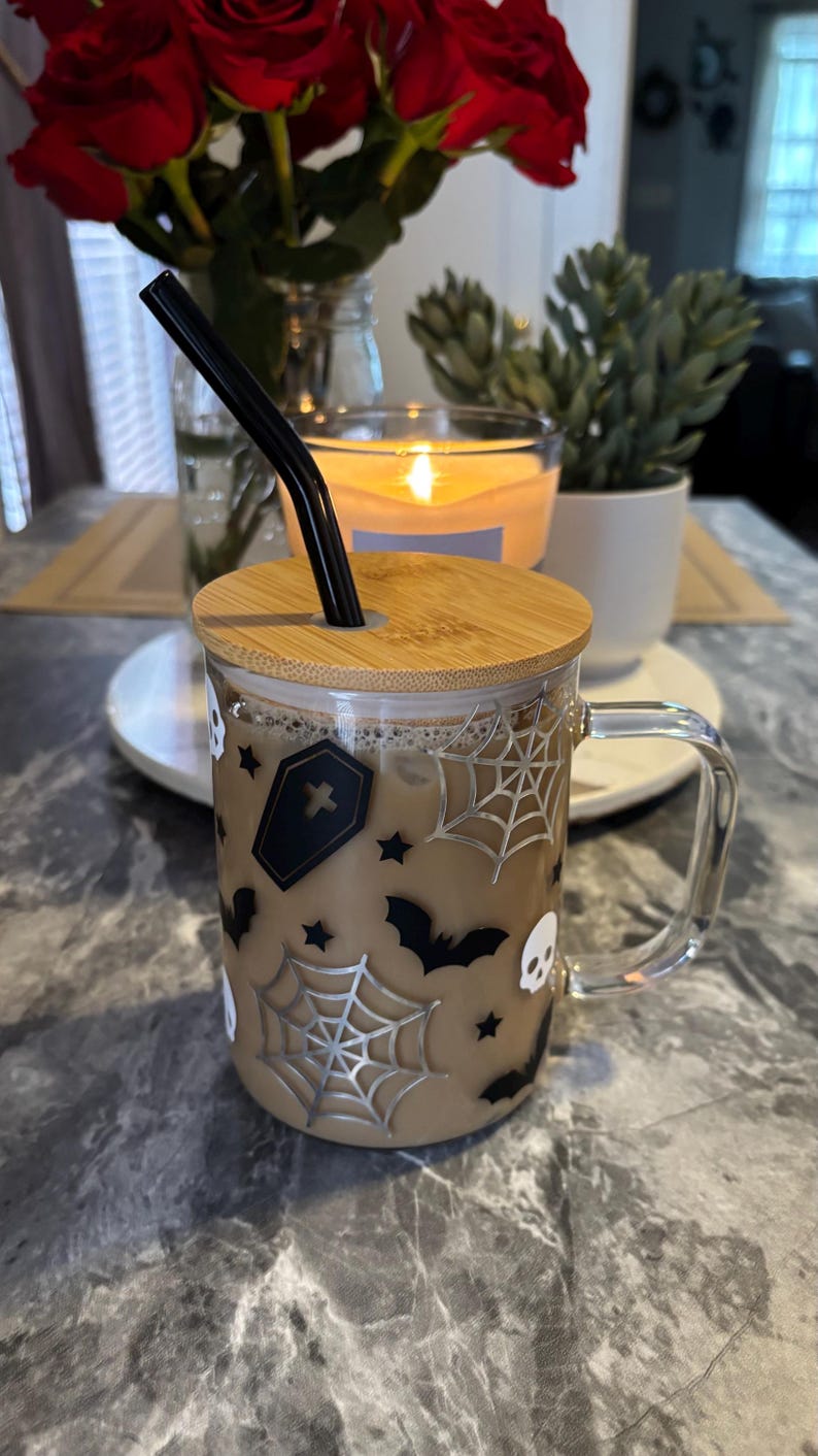 Pode incluir: Uma caneca de vidro com um design de Halloween marrom e preto. A caneca tem uma tampa de bambu e um canudo preto. O design inclui morcegos, teias de aranha, estrelas e caveiras.