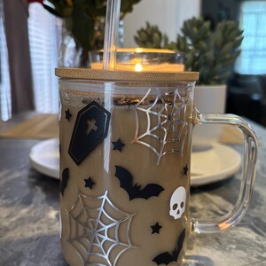 Pode incluir: Uma caneca de vidro com tampa de bambu e al&ccedil;a transparente. A caneca est&aacute; cheia de um l&iacute;quido marrom e decorada com desenhos tem&aacute;ticos de Halloween em preto e prata, incluindo morcegos, teias de aranha e um caix&atilde;o.