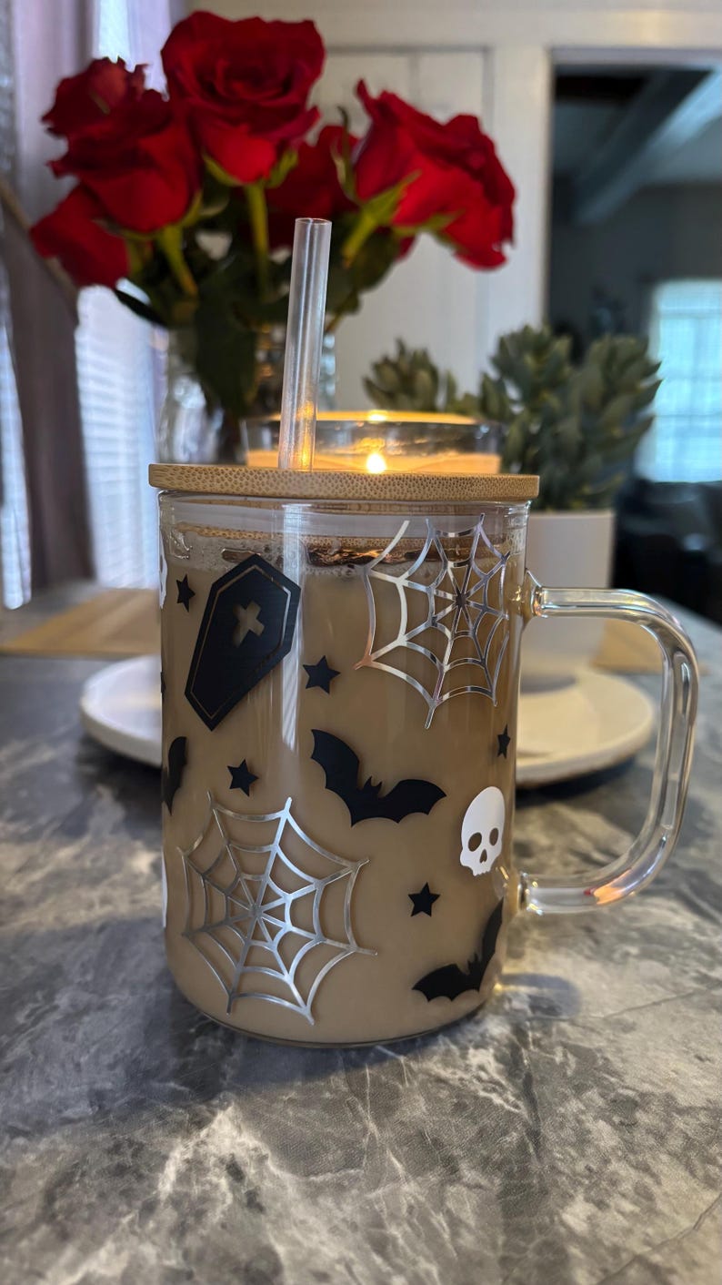 Pode incluir: Uma caneca de vidro com tampa de bambu e al&ccedil;a transparente. A caneca est&aacute; cheia de um l&iacute;quido marrom e decorada com desenhos tem&aacute;ticos de Halloween em preto e prata, incluindo morcegos, teias de aranha e um caix&atilde;o.