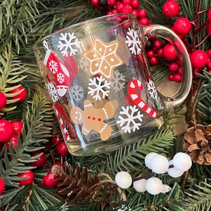 May include: A clear glass mug with a white handle decorated with a Christmas theme. The mug features gingerbread men, candy canes, snowflakes, and stars. The mug is surrounded by a green garland with red berries.