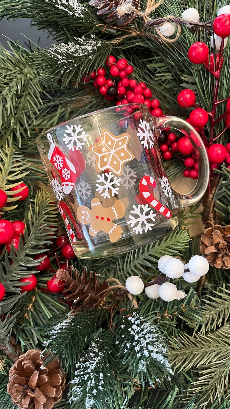 May include: A clear glass mug with a white handle decorated with a Christmas theme. The mug features gingerbread men, candy canes, snowflakes, and stars. The mug is surrounded by a green garland with red berries.