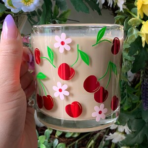 May include: A glass mug with a cream-colored interior and a cherry and flower design. The mug is filled with a light brown beverage.