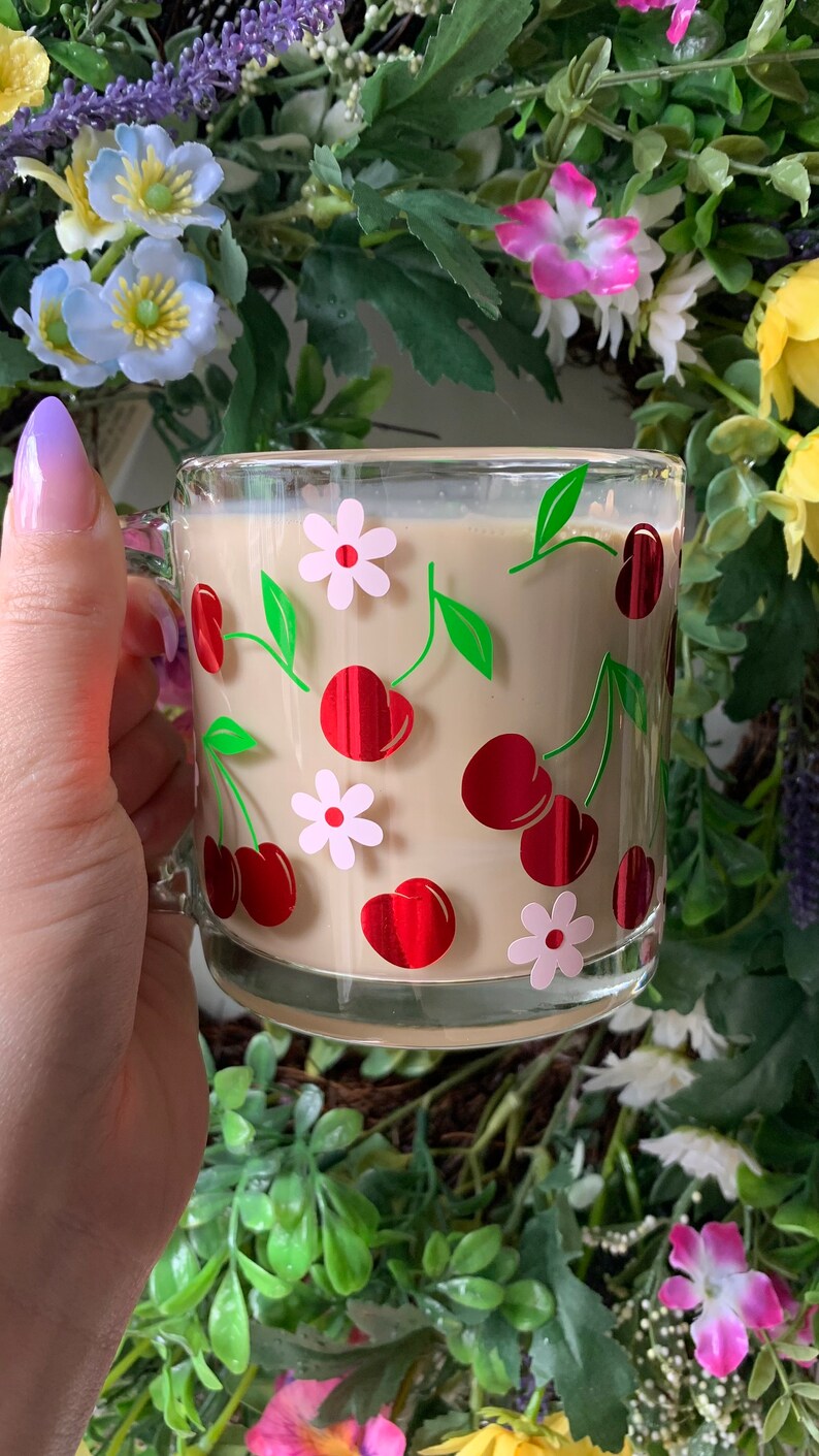 May include: A glass mug with a cream-colored interior and a cherry and flower design. The mug is filled with a light brown beverage.