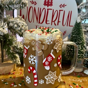 May include: A glass mug with a brown drink topped with whipped cream and sprinkles. The mug has a Christmas theme with gingerbread men, snowflakes, candy canes, and stockings.