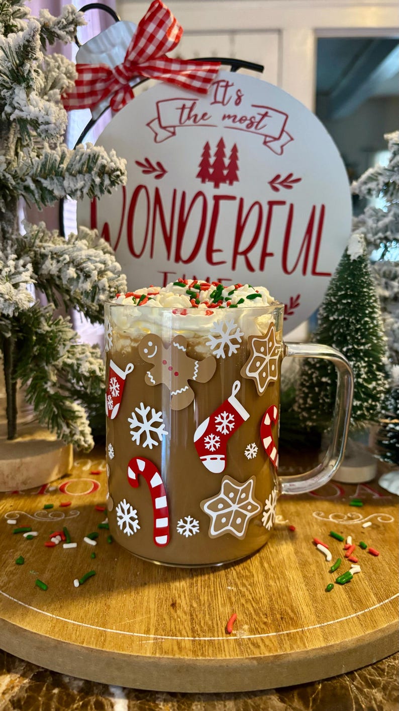 May include: A glass mug with a brown drink topped with whipped cream and sprinkles. The mug has a Christmas theme with gingerbread men, snowflakes, candy canes, and stockings.
