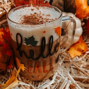 Op de afbeelding: Een glazen mok met een wit en bruin drankje erin. De mok heeft de tekst "hello fall" erop geschreven in het zwart. De mok staat op een bed van stro met herfstbladeren en pompoenen eromheen.