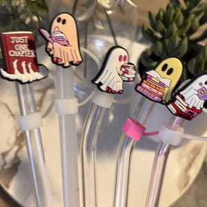Puede incluir: Cinco pajitas de vidrio transparente con adornos de personajes de fantasmas y libros coloridos. Los adornos est&aacute;n unidos a las pajitas con anillos de pl&aacute;stico blanco. Las pajitas se sostienen en una mano. El libro de un adorno dice "Just One Chapter".