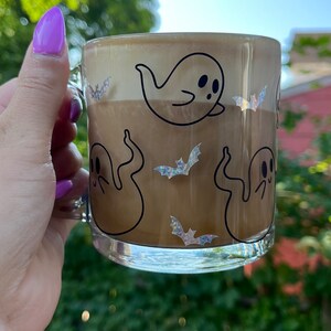 May include: A clear glass mug with a brown beverage inside. The mug has a black and white ghost and bat design.