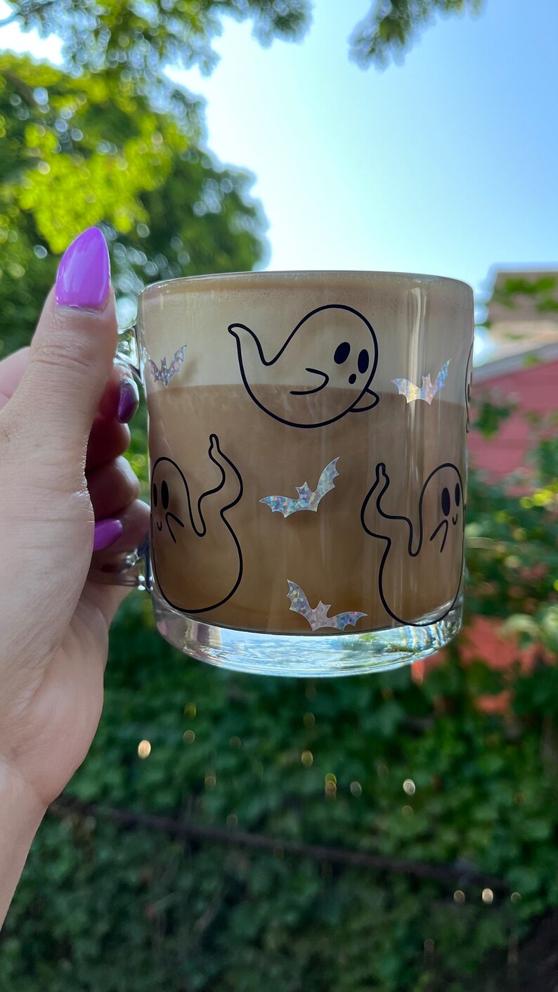 May include: A clear glass mug with a brown beverage inside. The mug has a black and white ghost and bat design.