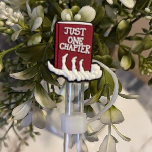 Puede incluir: Un charm de libro rojo con texto blanco que dice "JUST ONE CHAPTER" unido a una pajita de pl&aacute;stico transparente. El charm tiene forma de mano sosteniendo el libro.
