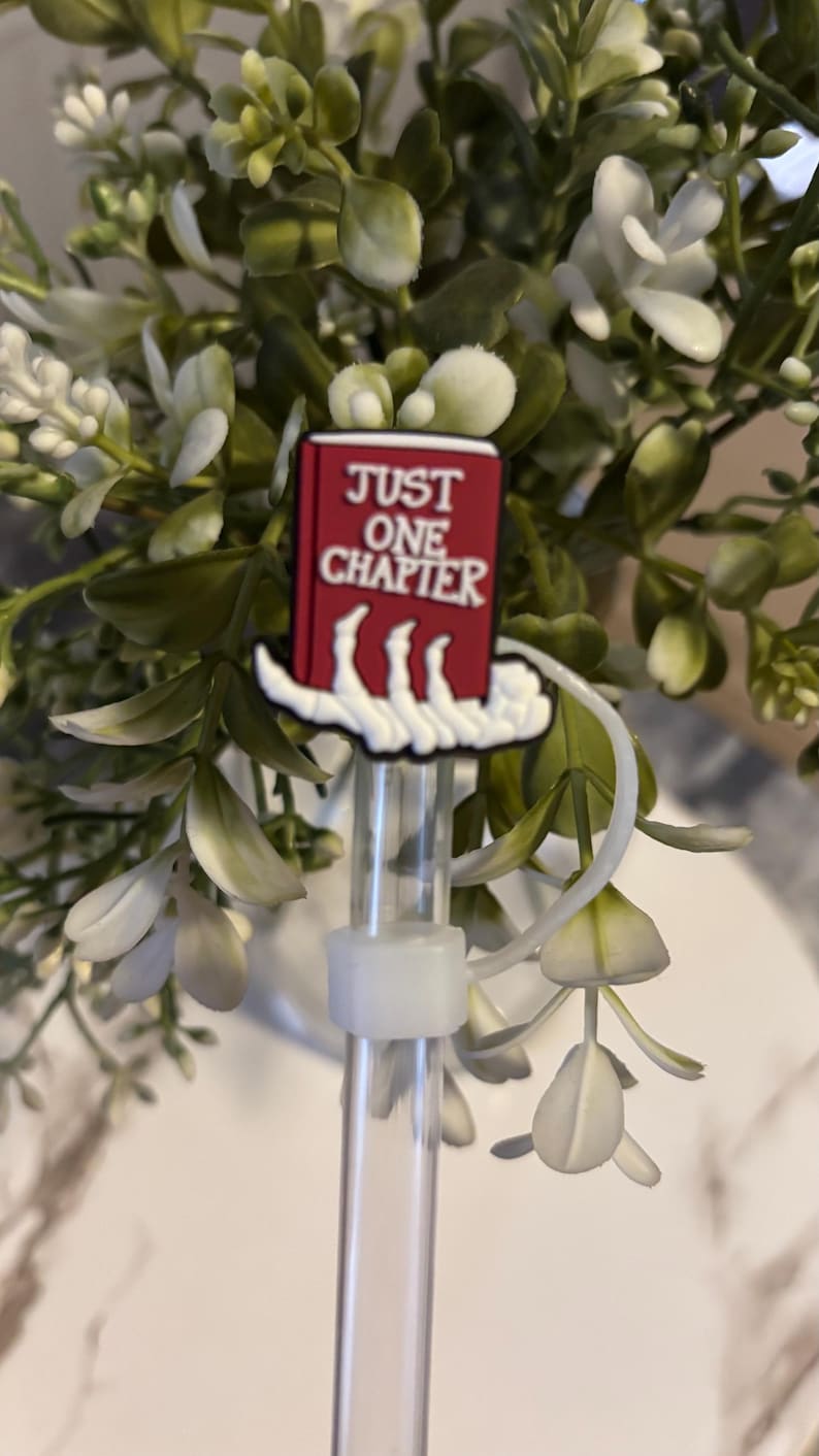 Puede incluir: Un charm de libro rojo con texto blanco que dice "JUST ONE CHAPTER" unido a una pajita de pl&aacute;stico transparente. El charm tiene forma de mano sosteniendo el libro.