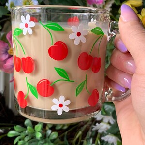 May include: A glass mug with a handle, featuring a pattern of red cherries and white flowers on a beige background. The mug is filled with a light brown liquid.