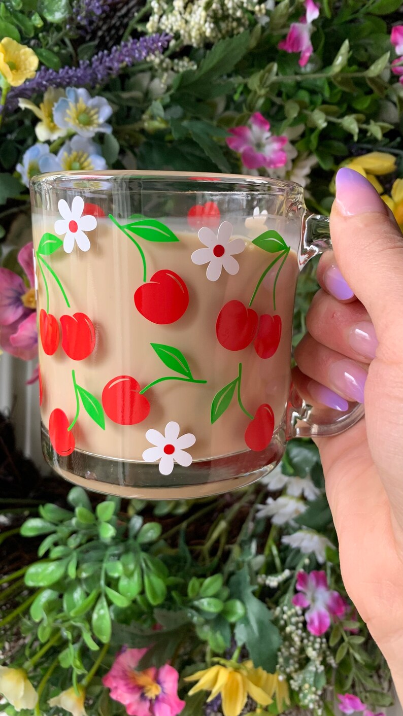 May include: A glass mug with a handle, featuring a pattern of red cherries and white flowers on a beige background. The mug is filled with a light brown liquid.