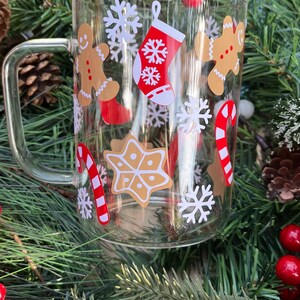May include: A clear glass mug with a handle decorated with Christmas-themed stickers. The stickers feature gingerbread men, candy canes, snowflakes, and a stocking.