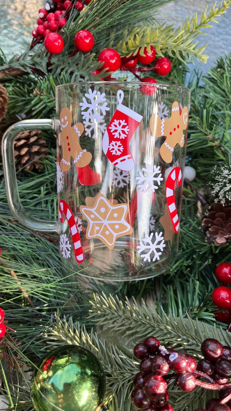 May include: A clear glass mug with a handle decorated with Christmas-themed stickers. The stickers feature gingerbread men, candy canes, snowflakes, and a stocking.