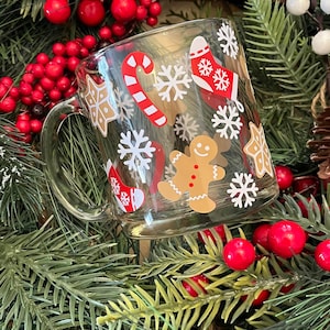 May include: A clear glass mug with a Christmas theme. The mug is decorated with white snowflakes, candy canes, gingerbread men, and stockings. The mug is surrounded by green pine branches and red berries.