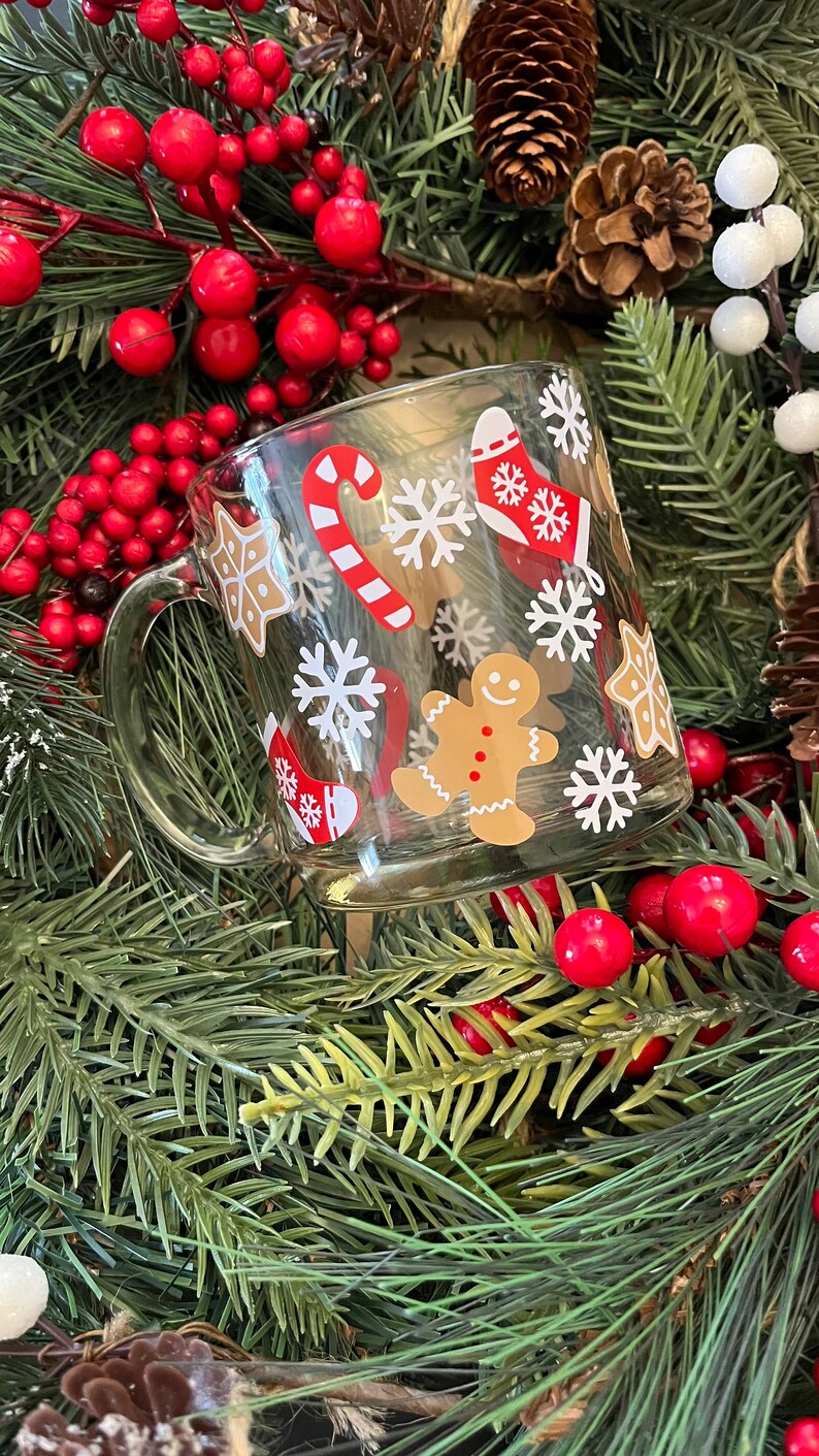 May include: A clear glass mug with a Christmas theme. The mug is decorated with white snowflakes, candy canes, gingerbread men, and stockings. The mug is surrounded by green pine branches and red berries.