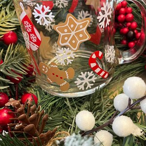 May include: A clear glass mug with a festive Christmas design. The mug features gingerbread men, candy canes, snowflakes, and stars. The mug is surrounded by a green garland with red berries and white balls.