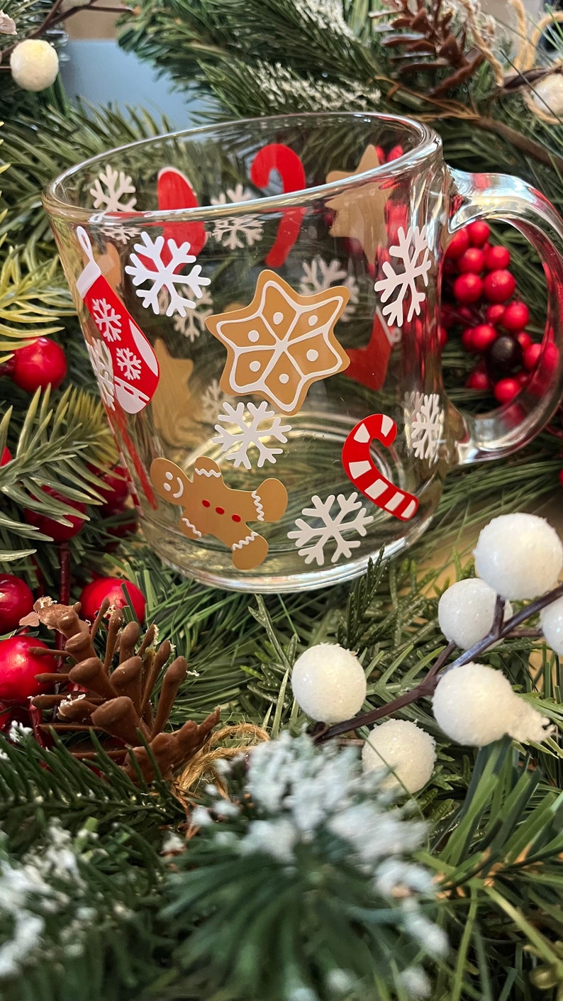 May include: A clear glass mug with a festive Christmas design. The mug features gingerbread men, candy canes, snowflakes, and stars. The mug is surrounded by a green garland with red berries and white balls.