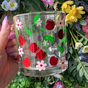 May include: A clear glass mug with a red and green cherry design. The mug has a white handle and is decorated with pink and red flowers.