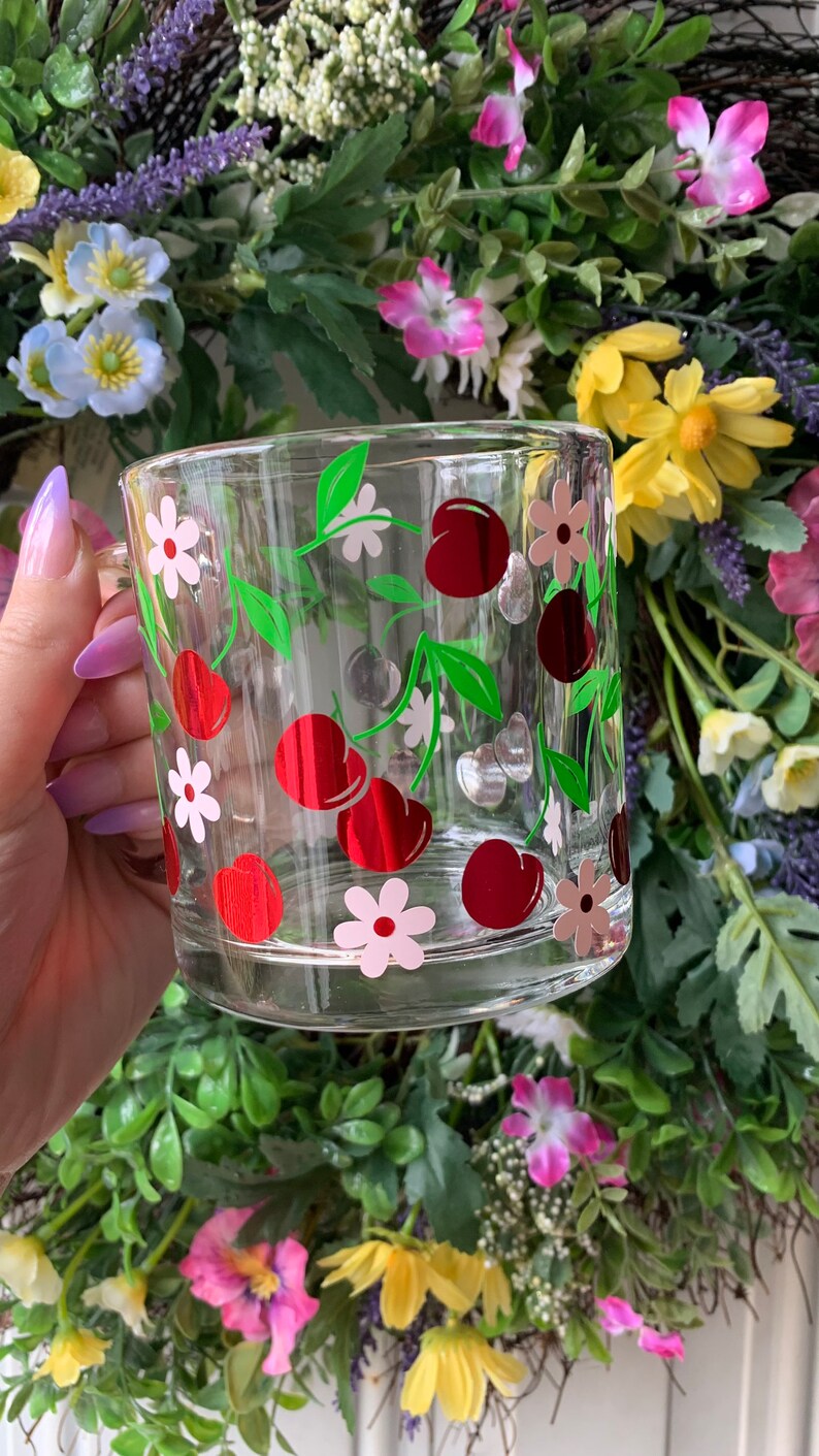 May include: A clear glass mug with a red and green cherry design. The mug has a white handle and is decorated with pink and red flowers.