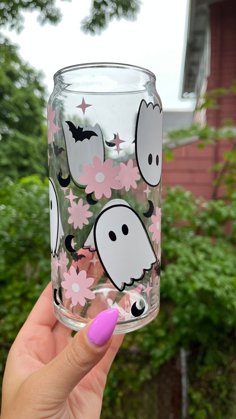 May include: A clear glass can with a pink and white ghost, flower, and crescent moon design. The can is decorated with a black bat and stars.