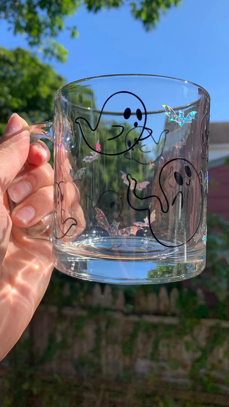 May include: A clear glass mug with a black outline of ghosts and bats. The ghosts have two black dots for eyes. The bats have iridescent glitter.