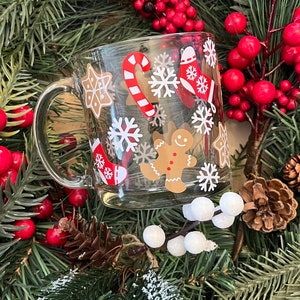May include: A clear glass mug with a red and white candy cane, gingerbread man, snowflakes, and stars design. The mug is surrounded by a green Christmas wreath with red berries and pine cones.