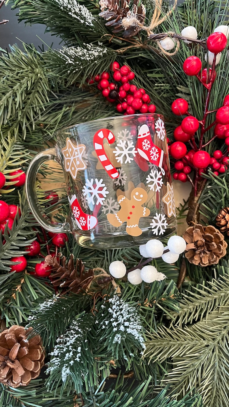 May include: A clear glass mug with a red and white candy cane, gingerbread man, snowflakes, and stars design. The mug is surrounded by a green Christmas wreath with red berries and pine cones.