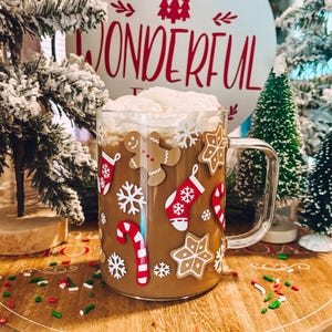 May include: A glass mug with a festive Christmas design featuring gingerbread men, snowflakes, candy canes, and stockings. The mug is filled with a brown beverage topped with whipped cream. The mug is sitting on a wooden tray with red and green sprinkles.