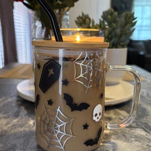 Pode incluir: Uma caneca de vidro transparente com tampa de bambu e um canudo preto. A caneca tem um tema de Halloween com morcegos pretos, teias de aranha e um design de caveira. A caneca est&aacute; cheia de um l&iacute;quido castanho.