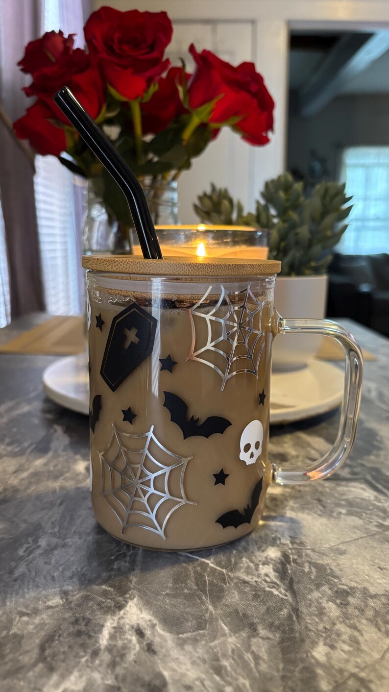 Pode incluir: Uma caneca de vidro transparente com tampa de bambu e um canudo preto. A caneca tem um tema de Halloween com morcegos pretos, teias de aranha e um design de caveira. A caneca est&aacute; cheia de um l&iacute;quido castanho.