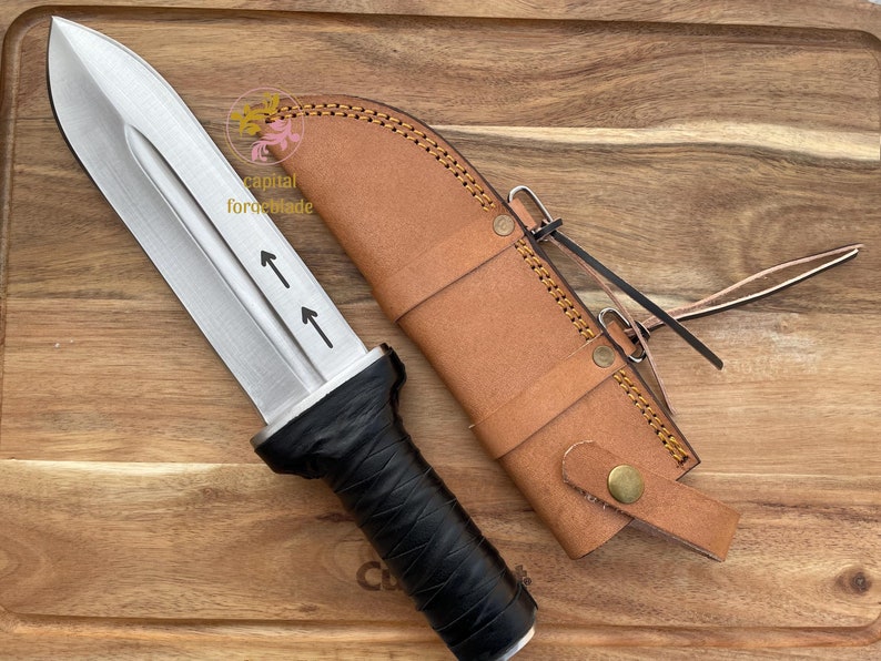 Pu&ograve; includere: Un coltello argentato con manico nero e fodero in pelle. Il coltello ha due frecce che puntano verso l'alto sulla lama. Il fodero ha una cinghia in pelle marrone con una fibbia in ottone.