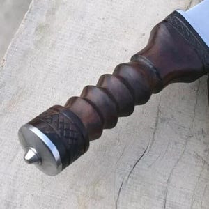 Pu&ograve; includere: Primo piano di un coltello fatto a mano con un manico in legno marrone scuro a spirale e un'estremit&agrave; in metallo argentato. La lama &egrave; parzialmente visibile, cos&igrave; come un fodero in pelle marrone con cuciture decorative.