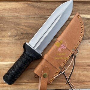 Pu&ograve; includere: Un coltello argentato con manico nero e fodero in pelle marrone. Il fodero ha un design cucito e una cinghia con fibbia.