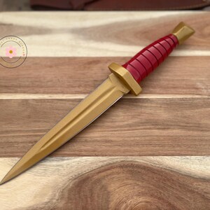 Peut inclure: Un poignard d&eacute;coratif avec une lame et une garde dor&eacute;es, et un manche rouge. Le poignard est pr&eacute;sent&eacute; sur une planche &agrave; d&eacute;couper en bois. La poign&eacute;e a un design nervur&eacute;. Le poignard est un objet d&eacute;coratif.
