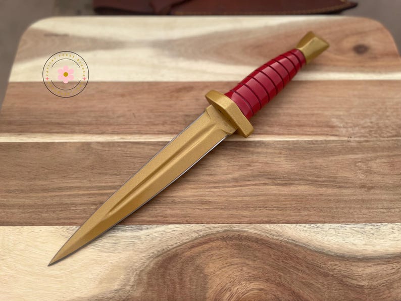 Peut inclure: Un poignard d&eacute;coratif avec une lame et une garde dor&eacute;es, et un manche rouge. Le poignard est pr&eacute;sent&eacute; sur une planche &agrave; d&eacute;couper en bois. La poign&eacute;e a un design nervur&eacute;. Le poignard est un objet d&eacute;coratif.